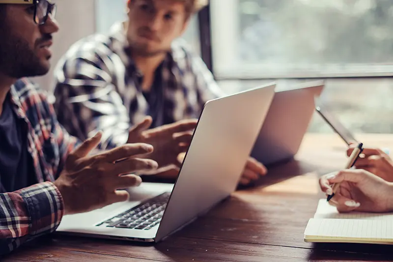 Two men sitting at a desk, talking. They both have laptops in front of them.