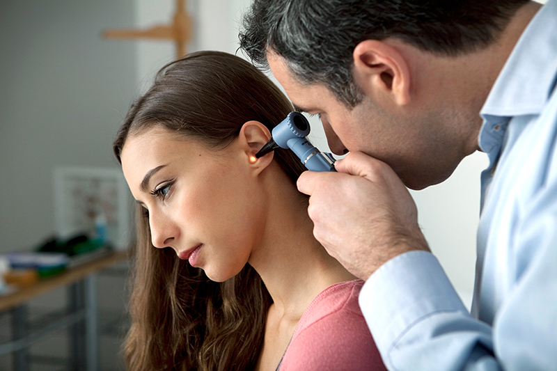 Woman sitting on a table while a doctor inspects her left ear with an otoscope